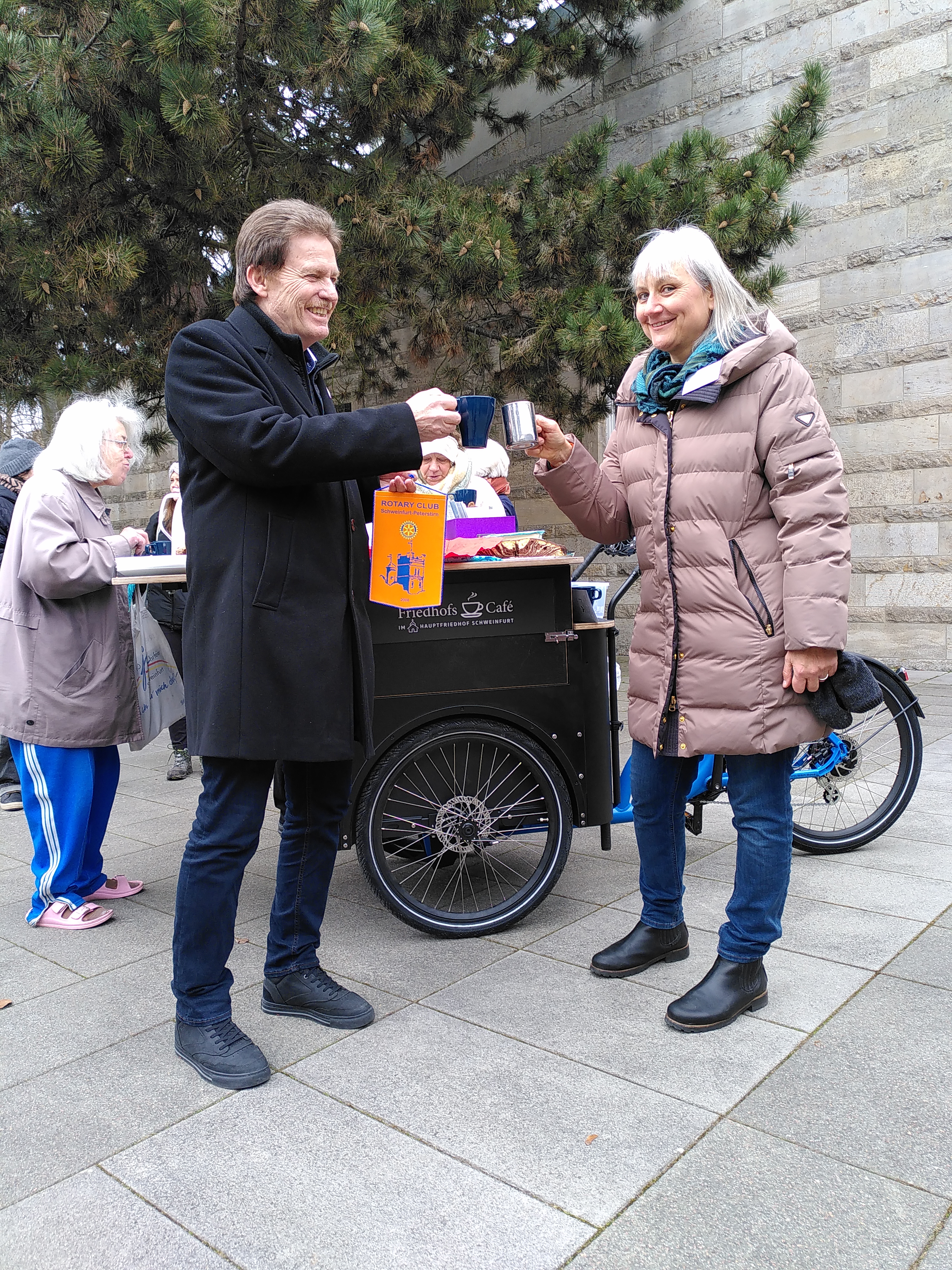 Ein Mann mit dunklem Mantel und eine grauhaarige Frau mit heller Jacke stoßen mit Kaffeebechern an. Der Mann übergibt einen orangen Club-Wimpel mit Rotary-Rad und einer Grafik darauf.